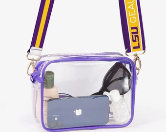 Könnte beinhalten: Transparente Umhängetasche mit lila Besatz und einem Riemen mit dem Text "LSU GEAUX TIGERS". Die Tasche enthält ein Telefon, eine Sonnenbrille, Händedesinfektionsmittel und andere persönliche Gegenstände. Der Riemen ist lila mit einem gelben Streifen.