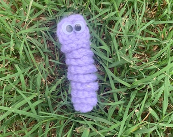 May include: A purple crocheted worm with googly eyes. The worm is lying on a bed of green grass.
