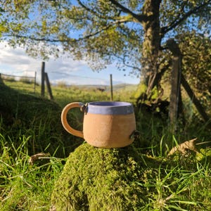 May include: A handcrafted ceramic mug with a light brown exterior and a lavender rim. The mug features a curved handle and small decorative details. It is set on green moss, with a natural outdoor background of grass and trees.