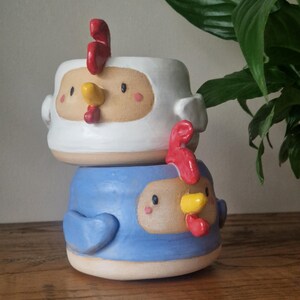 May include: Two ceramic planters shaped like chickens. The top planter is white with a red comb and a yellow beak. The bottom planter is blue with a red comb and a yellow beak.