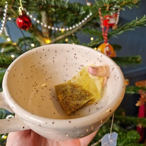 May include: A white speckled ceramic mug containing a tea bag. The mug is held in front of a Christmas tree decorated with red baubles. The tea bag is yellow with a pink tag.