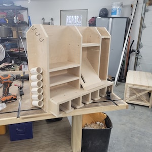 May include: A wooden storage unit with shelves, a drawer, and a slanted compartment. The unit is made of plywood and has a light brown finish. It is sitting on a workbench.