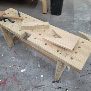 May include: A wooden workbench with a light brown finish, featuring a series of evenly spaced holes. A wooden mallet and a metal clamp rest on the bench. Additional wooden pieces, including a triangular shape, are also present.