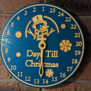 May include: A blue wooden countdown clock with a snowman design and a yellow hand pointing to the number 15. The clock reads "Days Till Christmas".