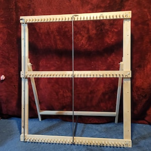 May include: A wooden weaving loom with a rectangular frame and numerous pegs along the top and bottom edges. The loom has a stand with angled legs for support. A metal rod runs vertically through the centre. The loom is made of light-coloured wood.