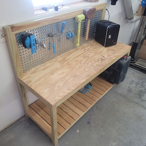 Może przedstawiać: Drewniany stół warsztatowy z metalową tablicą perforowaną z tyłu. Stół warsztatowy ma drewniany blat, dolną półkę i mały czarny głośnik na górze. Na tablicy perforowanej wiszą różne narzędzia, w tym młotek, klucz i szczypce.