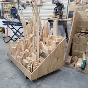May include: A wooden storage rack with various sizes of wood pieces organised inside. The rack is on wheels and has a slanted design. The rack is in a workshop setting with other wood pieces and tools visible.