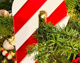 May include: A red and white candy cane ornament hanging on a Christmas tree. The candy cane is made of wood and has a white stripe down the middle.