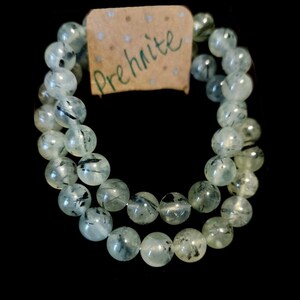 May include: A green prehnite bracelet with black inclusions. The bracelet is made of round beads and is strung on a clear elastic cord. The bracelet is labelled "Prehnite".