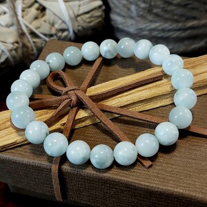 May include: A light blue gemstone bracelet with a brown leather tie closure. The bracelet is on a brown box with a wooden stick inside.