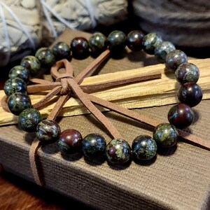 May include: A green and brown gemstone bracelet with a brown leather cord tie. The bracelet is made of round beads and is sitting on a brown box.