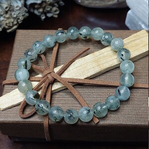 May include: A green and black gemstone bracelet with a brown leather cord tied around it. The bracelet is in a brown box with a wooden stick inside.