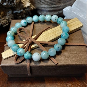May include: A teal green gemstone bracelet with brown leather ties, displayed in a brown gift box with a wooden stick inside.