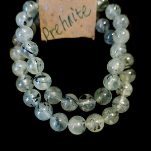 May include: A double-strand bracelet made of green prehnite beads with black inclusions. The bracelet has a brown paper tag with the word "Prehnite" written in green.