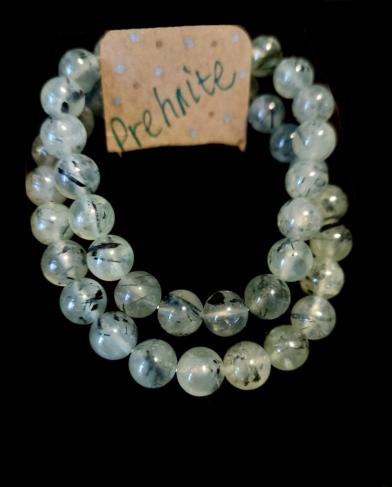 May include: A double-strand bracelet made of green prehnite beads with black inclusions. The bracelet has a brown paper tag with the word "Prehnite" written in green.