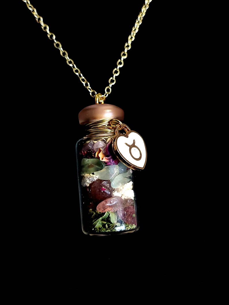 Peut inclure: Un collier &agrave; cha&icirc;ne dor&eacute;e avec un petit pendentif en forme de flacon en verre. Le flacon est rempli de cristaux color&eacute;s, de fleurs s&eacute;ch&eacute;es et d'herbes. Un charm en forme de c&oelig;ur dor&eacute; avec un symbole du Taureau est suspendu.