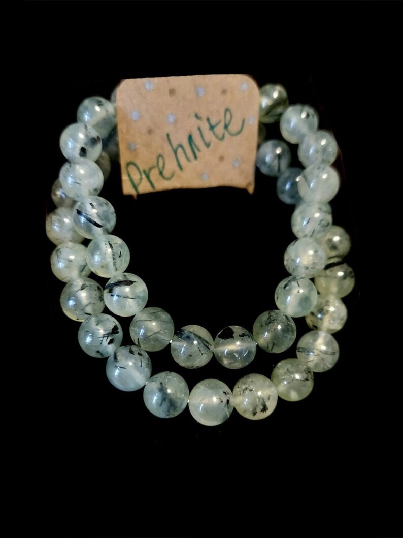 May include: A green prehnite bracelet with black inclusions. The bracelet is made of round beads and is strung on a clear elastic cord. The bracelet is labelled "Prehnite".