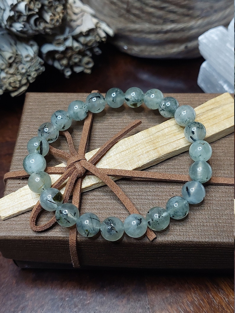 May include: A green and black gemstone bracelet with a brown leather cord tied around it. The bracelet is in a brown box with a wooden stick inside.