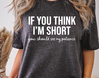May include: Dark gray t-shirt with the text "IF YOU THINK I'M SHORT you should see my patience" in white. The shirt is worn with distressed blue jeans. The image is taken indoors.