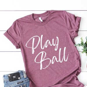 May include: A maroon t-shirt with the words "Play Ball" written in white on the front. The shirt is laying on a white wooden surface with a pair of white sneakers and a pair of blue jeans.