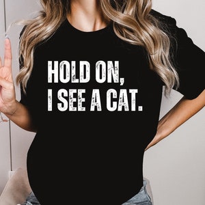 May include: A black t-shirt with the text "Hold on, I see a cat." printed in white. The person wearing the shirt is also wearing ripped blue jeans.
