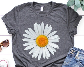 May include: A dark gray t-shirt with a white daisy graphic. The daisy has a yellow center and white petals. The t-shirt is laid flat on a white surface with a pair of white sneakers, blue jeans, and sunglasses.