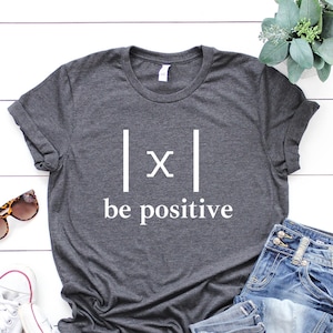 May include: A dark gray t-shirt with a white graphic that says "|x| be positive". The shirt is laid flat on a white wooden surface with a pair of sunglasses, a pair of white sneakers, a pair of blue jeans, and a potted plant.
