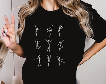 May include: Black t-shirt with a graphic of skeletons doing ballet poses. The shirt is being worn by a person with long blonde hair and blue jeans.