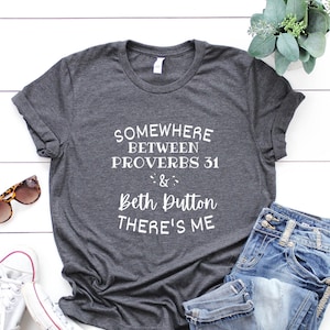 May include: Dark gray t-shirt with white text that reads "Somewhere Between Proverbs 31 & Beth Dutton THERE'S ME." The shirt is paired with blue jeans, white sneakers, and sunglasses. A sprig of greenery is also present.