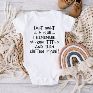 May include: White baby onesie with black text that reads "LAST NIGHT IS A BLUR... I REMEMBER SUCKING TITTIES AND THEN SHITTING MYSELF." The onesie is on a woven basket with a rainbow decoration and baby shoes.