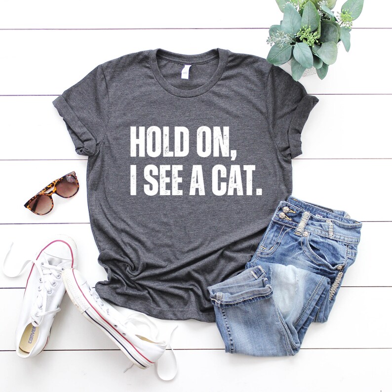 May include: A dark gray t-shirt with the text "HOLD ON, I SEE A CAT." printed in white. The shirt is laid flat on a white wooden surface with a pair of white sneakers, blue jeans, and sunglasses.