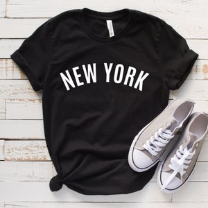 May include: Black t-shirt with white text that reads "NEW YORK". The shirt is laid flat on a white wooden surface with a pair of gray canvas shoes.
