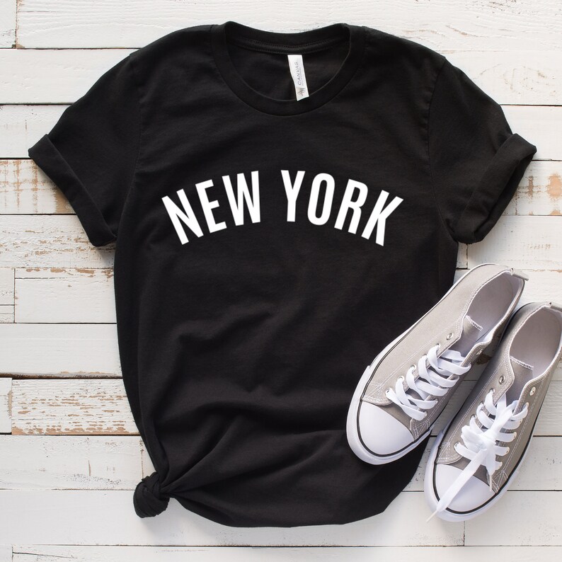 May include: Black t-shirt with white text that reads "NEW YORK". The shirt is laid flat on a white wooden surface with a pair of gray canvas shoes.