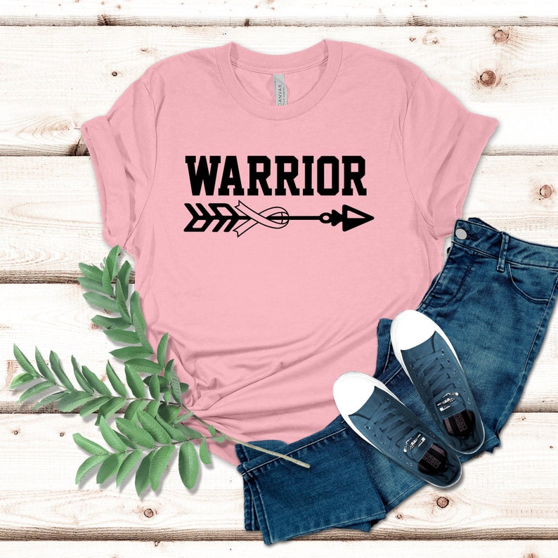 Puede incluir: Camiseta rosa con un dise&ntilde;o de flecha y cinta negra. El texto "WARRIOR" est&aacute; impreso en negro sobre la flecha.