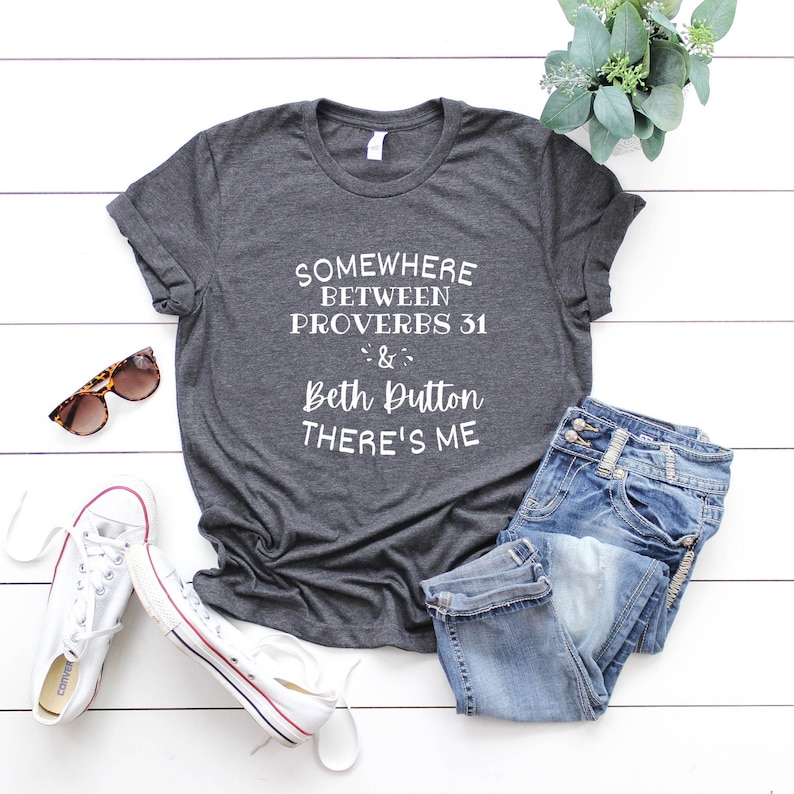 May include: A dark charcoal gray short-sleeve t-shirt featuring white text that reads: SOMEWHERE BETWEEN PROVERBS 31 & BETH DUTTON THERE'S ME. The shirt is displayed alongside blue denim jeans, white canvas sneakers, and tortoise shell sunglasses.