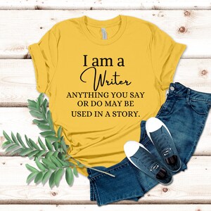 May include: A yellow t-shirt with the text "I am a Writer ANYTHING YOU SAY OR DO MAY BE USED IN A STORY."
