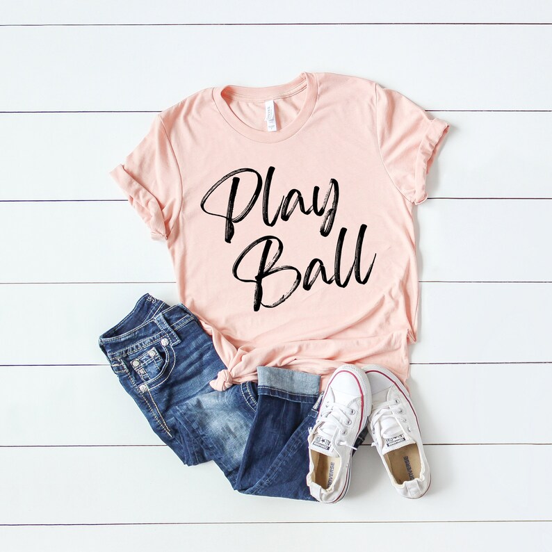 May include: A peach-colored t-shirt with the text "Play Ball" written in black script.