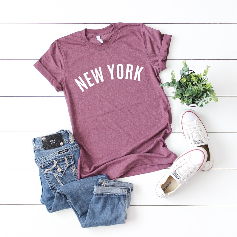 May include: A maroon t-shirt with the text "NEW YORK" in white lettering. The shirt is laid out on a white wooden surface with a pair of white sneakers and blue jeans.