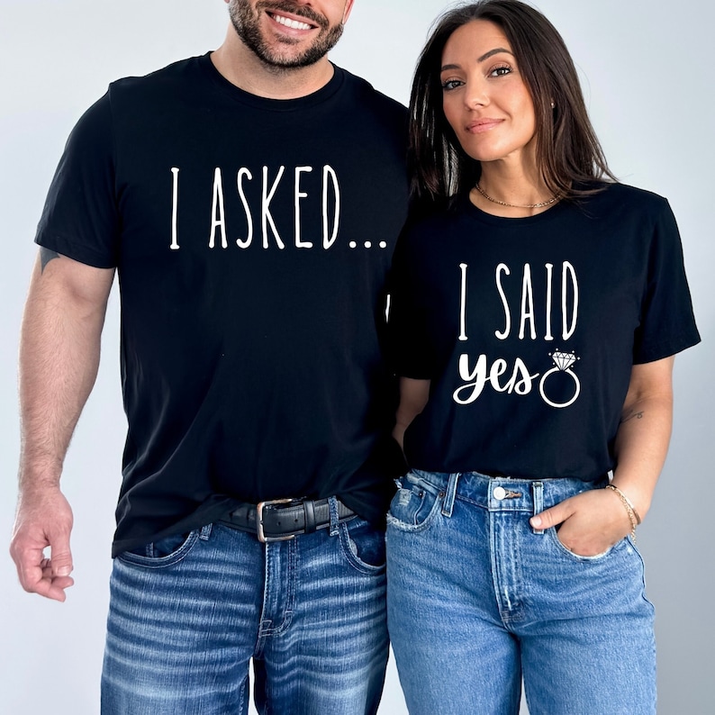 K&ouml;nnte beinhalten: Ein Paar tr&auml;gt passende schwarze T-Shirts mit wei&szlig;em Text. Das Shirt des Mannes sagt "Ich habe gefragt..." und das Shirt der Frau sagt "Ich habe ja gesagt" mit einer Ringgrafik.