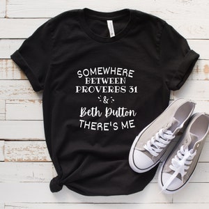 May include: A black short-sleeved t-shirt with white text that reads: SOMEWHERE BETWEEN PROVERBS 31 & Beth Dutton THERE'S ME. The shirt features a crew neckline and rolled sleeves, paired with a pair of grey low-top canvas sneakers with white laces.