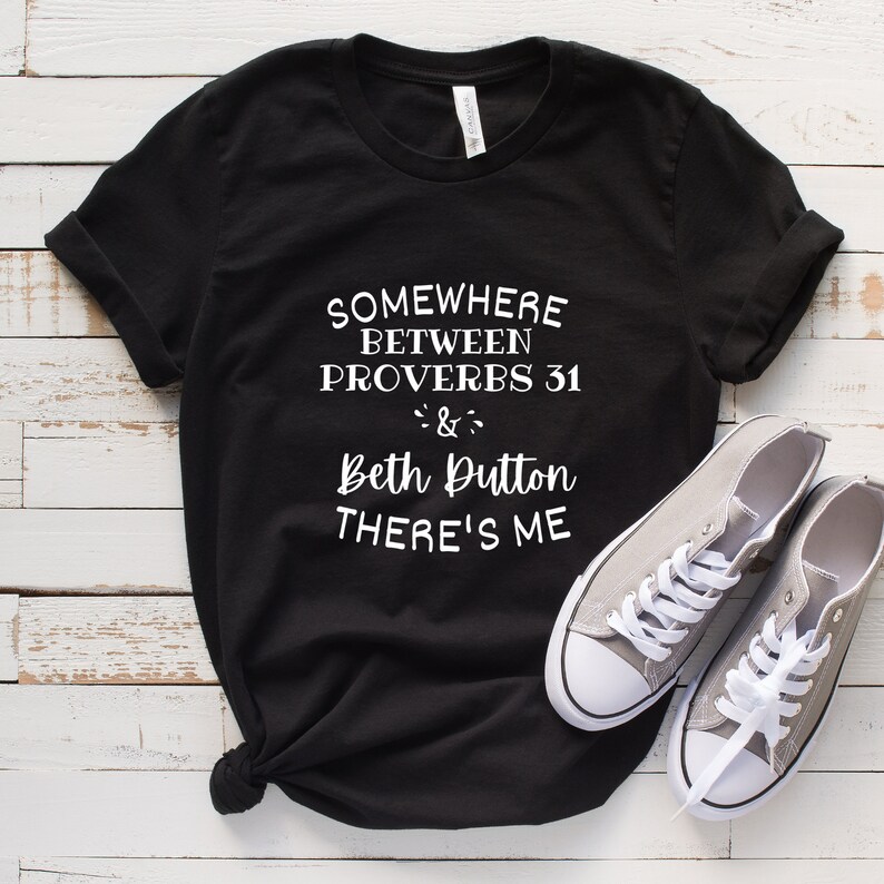 May include: A black short-sleeved t-shirt with white text that reads: SOMEWHERE BETWEEN PROVERBS 31 & Beth Dutton THERE'S ME. The shirt features a crew neckline and rolled sleeves, paired with a pair of grey low-top canvas sneakers with white laces.