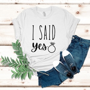 K&ouml;nnte beinhalten: Wei&szlig;es T-Shirt mit schwarzem Text, der "Ich habe Ja gesagt" lautet, mit einer Diamant-Ring-Grafik.