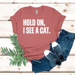 May include: A rust-colored t-shirt with the text "HOLD ON, I SEE A CAT." printed in white.