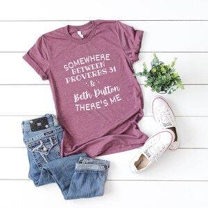 May include: A heathered maroon short-sleeve t-shirt with white text that reads: SOMEWHERE BETWEEN PROVERBS 31 & Beth Dutton THERE'S ME. The shirt is paired with blue denim jeans featuring a black leather patch and white canvas sneakers.