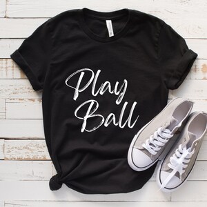 May include: Black t-shirt with white text that says "Play Ball". The shirt is laying on a white wooden surface with two gray canvas shoes.