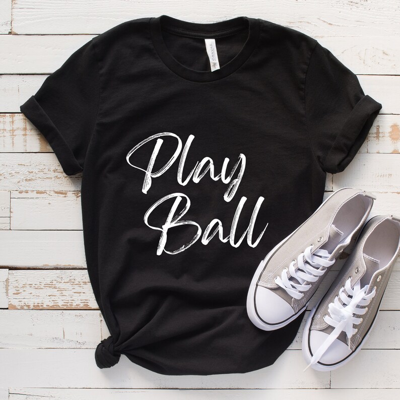 May include: Black t-shirt with white text that says "Play Ball". The shirt is laying on a white wooden surface with two gray canvas shoes.