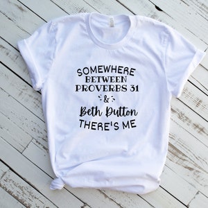 May include: A white short-sleeved t-shirt with black text printed on the front that reads: SOMEWHERE BETWEEN PROVERBS 31 & BETH DUTTON THERE'S ME. The shirt features a crew neckline and is displayed with the bottom hem tied in a small knot.