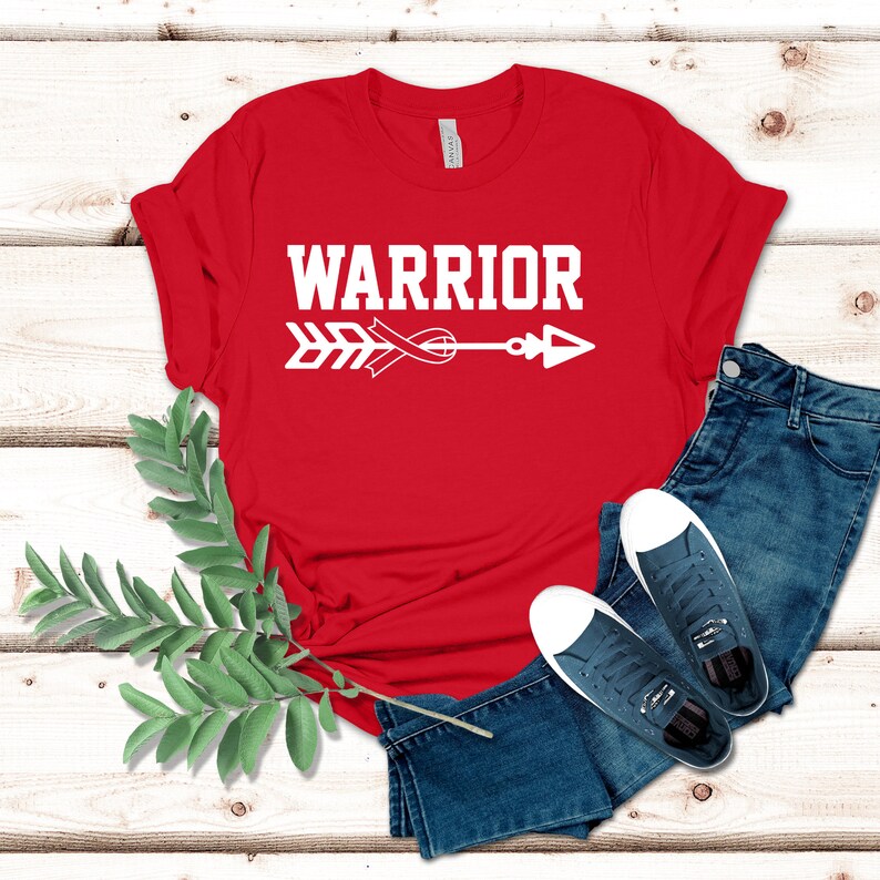 Puede incluir: Camiseta roja con texto blanco que dice "WARRIOR" y una flecha blanca con una cinta roja en el medio.