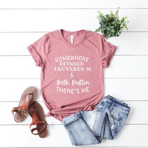 May include: A mauve short-sleeved t-shirt with white text that reads "SOMEWHERE BETWEEN PROVERBS 31 & BETH DUTTON THERE'S ME". The shirt is paired with blue denim jeans and brown sandals, with a small potted green plant nearby.