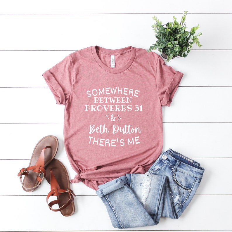 May include: A mauve short-sleeved t-shirt with white text that reads "SOMEWHERE BETWEEN PROVERBS 31 & BETH DUTTON THERE'S ME". The shirt is paired with blue denim jeans and brown sandals, with a small potted green plant nearby.